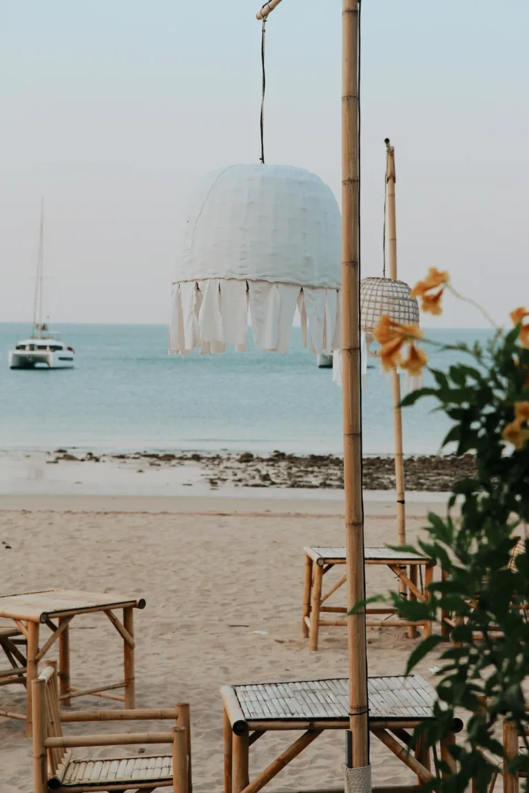 Vertical lifestyle photography of a beachfront lounge with a bohemian fringe lamp, bamboo furniture, and a catamaran anchored in the sea at sunset.